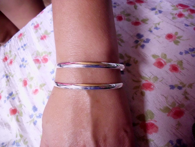 May include: Two silver bangles on a person's wrist. The bangles are simple and have a smooth, polished finish.