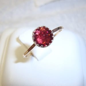 Pode incluir: Um anel de prata com uma gema vermelha engastada em uma moldura em forma de coroa.