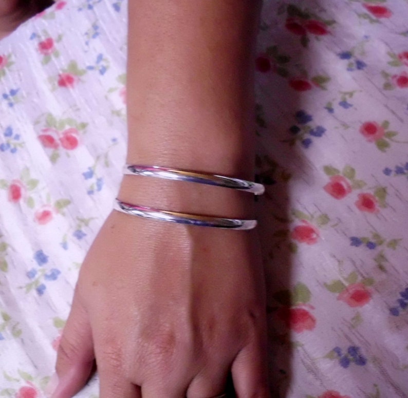 May include: A silver bangle bracelet with a polished finish. The bracelet is worn on a person's wrist.