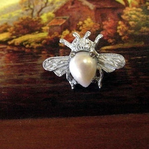 Puede incluir: Un broche de abeja de plata con un centro de perla. La abeja tiene alas detalladas y una corona en la cabeza.