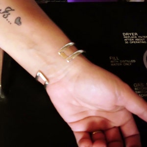 May include: A close-up of a person's wrist wearing a silver bangle bracelet. The person has a tattoo on their forearm that says "Here's..." with a small heart.