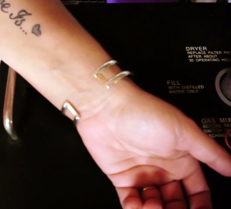 May include: A close-up of a person's wrist wearing a silver bangle bracelet. The person has a tattoo on their forearm that says "Here's..." with a small heart.