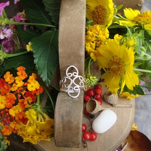 Puede incluir: Un anillo de plata con un diseño delicado e intrincado. El anillo está colocado sobre un fondo de flores coloridas, incluyendo girasoles amarillos y flores rosas y naranjas.