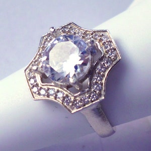 May include: A silver ring with a large, clear, round gemstone surrounded by smaller, clear gemstones. The ring has a decorative, geometric design.