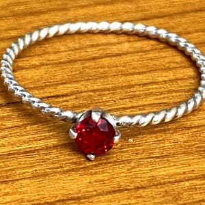 May include: A silver-toned ring with a twisted band and a vibrant red gemstone. The gemstone is round and faceted, set in a simple, four-prong setting. The ring is displayed on a wooden surface.