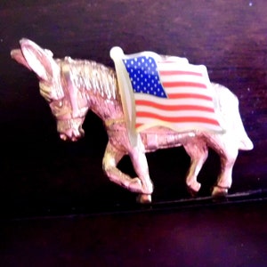 Puede incluir: Una figurita de burro de color dorado con una bandera estadounidense drapeada sobre su espalda. El burro está de pie sobre una superficie marrón oscura.