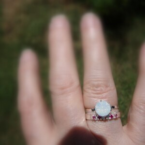 May include: A silver ring with a large, round, white opal stone. A smaller band with pink stones sits below the opal ring.