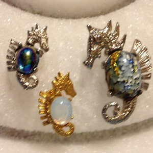 Puede incluir: Tres broches de caballito de mar de plata y oro con piedras de vidrio iridiscente y opalescente.