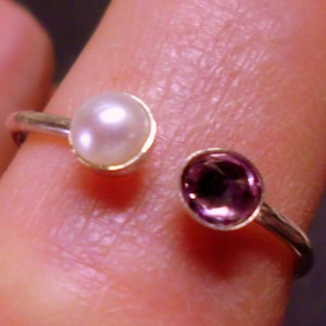 May include: A silver ring with a white pearl and a purple gemstone. The ring is on a finger.