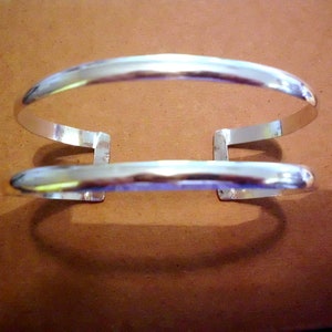 Könnte beinhalten: Ein silbernes Armreif-Armband mit einem Doppelband-Design. Das Armband ist offen und hat einen einfachen, minimalistischen Stil.