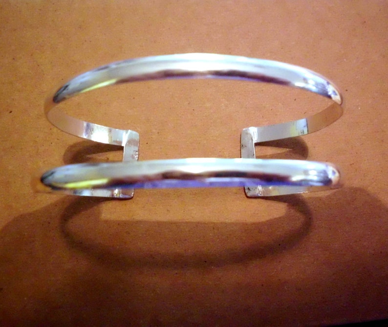 May include: A silver bangle bracelet with a double band design. The bracelet is open and has a simple, minimalist style.