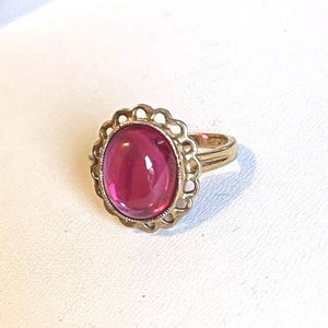 Może przedstawiać: Złoty pierścionek z dużym, owalnym, magentowym kamieniem. Kamień osadzony jest w dekoracyjnym, karbowanym obramowaniu. Obrączka pierścionka jest z gładkiego złota. Biżuteria w stylu vintage.