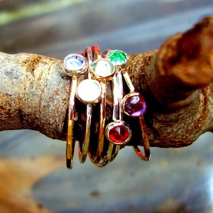 Op de afbeelding: Een set van zeven gouden ringen met gekleurde edelstenen. De ringen zijn op elkaar gestapeld en in een cirkel gerangschikt. De edelstenen zijn rood, groen, paars, wit en blauw.