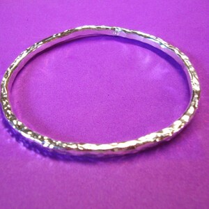 Puede incluir: Un brazalete de plata con textura martillada. El brazalete est&aacute; sobre un fondo morado.
