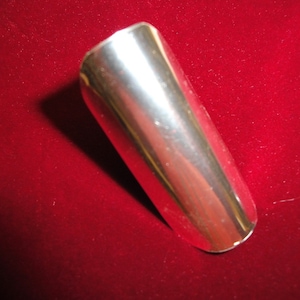 Puede incluir: Un protector de anillo de metal plateado, una cubierta protectora para un anillo, se muestra sobre un fondo de terciopelo rojo.