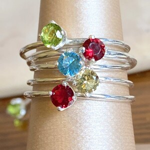 May include: Stack of five silver rings with colorful gemstones. The rings feature a green, red, blue, yellow, and red gemstone.