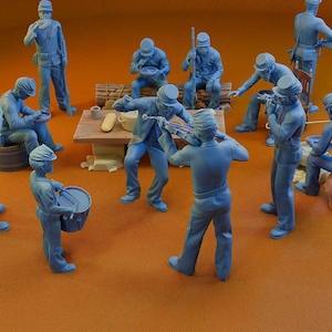 Op de afbeelding: Een verzameling blauwe miniatuurbeeldjes die een groep mensen in een kampeerscène uitbeelden. Sommige figuren zitten, andere staan, spelen instrumenten of eten. Een kleine tafel en vaten zijn ook aanwezig.