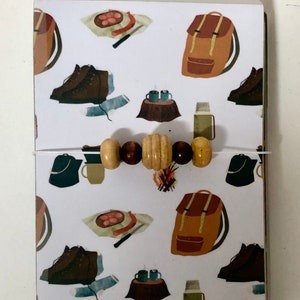 May include: A stack of notebooks with a white background and a repeating pattern of camping-themed illustrations. The illustrations include a backpack, hiking boots, a campfire, a thermos, and a plate of food. A string of wooden beads is wrapped around the notebooks.
