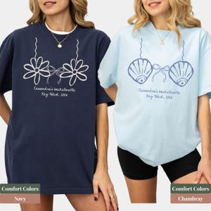 May include: Two t-shirts, one navy and one chambray, featuring a graphic design of a floral bra and seashell bra, respectively. The text "Cassandra's bachelorette Key West, 2016" is printed below the designs. The bottom of the image shows the color names.