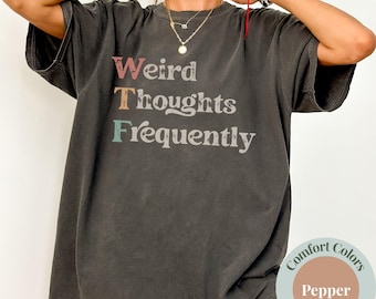 May include: Dark gray t-shirt with the text "Weird Thoughts Frequently" in a colorful, vintage-style font. The shirt is a relaxed fit and has short sleeves. The shirt is labeled "Comfort Colors" and the color is "Pepper".