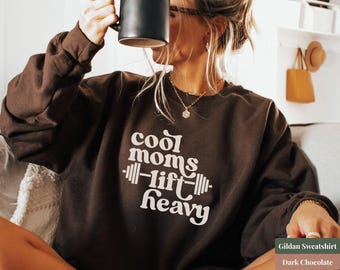 Könnte beinhalten: Dunkelbraunes Sweatshirt mit dem weißen Text "cool moms lift heavy". Das Sweatshirt hat einen Rundhalsausschnitt und lange Ärmel. Eine schwarze Tasse ist ebenfalls zu sehen.