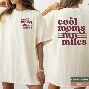 Puede incluir: Una camiseta de manga corta color marfil con el texto granate "cool moms run miles" impreso en una fuente retro en el pecho izquierdo y en un tamaño mayor en la espalda, con líneas horizontales que acentúan el diseño del texto.