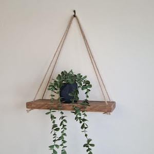 May include: A wooden hanging shelf with a dark grey geometric planter holding a green plant. The shelf is suspended by thick, natural-coloured rope from a metal hook. The shelf is made of reclaimed wood. The shelf is approximately 60 cm in length.