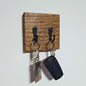 May include: A wooden key holder with two black metal hooks. The wood is stained a warm brown, showing the grain. Two sets of keys hang from the hooks, one with a Beatles keychain. A simple and functional home decor item.