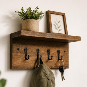 May include: A wall-mounted wooden shelf with a dark brown finish featuring four black metal hooks. A small potted green plant and a framed botanical print sit on the shelf. An olive green baseball cap hangs from one hook and keys hang from another.
