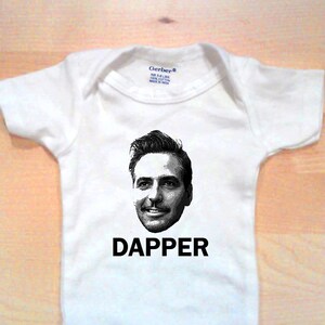 Op de afbeelding: Witte baby romper met een zwart-witte afbeelding van een mannengezicht en het woord "Dapper" in zwart gedrukt.