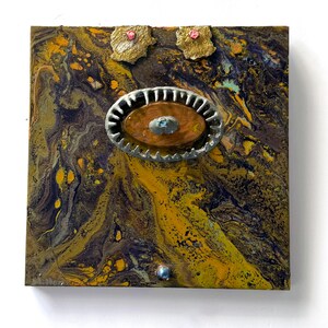 May include: Abstract artwork on a square wooden panel. The piece features a swirling pattern of yellow, brown, and purple hues. It is decorated with metal and jewel embellishments, including an oval-shaped centrepiece and two floral accents.