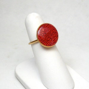 May include: A gold-toned ring featuring a large, round, red beaded design. The ring is displayed on a white finger-shaped stand, showcasing the vibrant color and texture of the jewelry.