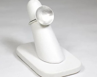 May include: A silver ring with a large, round, flat top. The ring is on a white ring display stand.