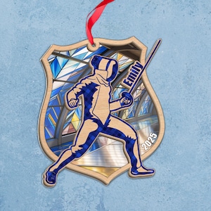 May include: Wooden shield-shaped ornament featuring a blue and tan fencing figure with the name "Emily" and the year "2025". The ornament has a red ribbon for hanging and a stained glass background.