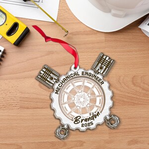 May include: A wooden ornament with a red ribbon for hanging. The ornament is shaped like a gear with the words "MECHANICAL ENGINEER" and the name "Brandon" and the year "2025" engraved on it. The ornament has a car wheel design in the center.