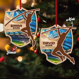May include: Two wooden Christmas ornaments with a shield shape, featuring a pole vaulter silhouette. One ornament says "EMMA 2025" and the other says "DAVID 2025". The background shows a stadium and stained glass design. Both ornaments have a red ribbon for hanging.