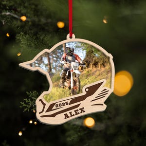 May include: Wooden Christmas ornament shaped like a motorcycle helmet, with a photo of a person riding a dirt bike. The ornament has the text "2025/ALEX" engraved on it and is suspended by a red ribbon.