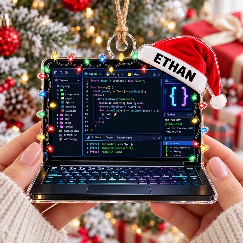 May include: A clear acrylic ornament shaped like a laptop displaying code, decorated with colorful string lights and a red Santa hat featuring the name ETHAN in black text. The ornament hangs from a jute twine loop and shows a software interface.