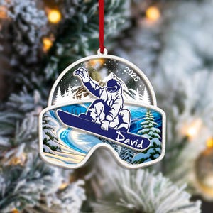 May include: A white and blue snowboarder ornament with the name "David" and the year "2025". The ornament features a snowboarder in action against a snowy mountain backdrop, with a red ribbon for hanging.