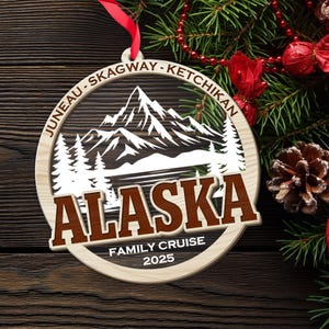 May include: Wooden Christmas ornament with a cut-out design featuring mountains, trees, and the word "ALASKA" in brown. The ornament also includes the text "JUNEAU - SKAGWAY - KETCHIKAN" and "FAMILY CRUISE 2025". A red ribbon is attached for hanging.