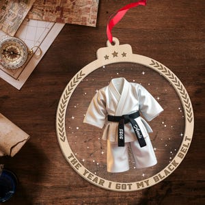 May include: Wooden ornament with a karate uniform design. The ornament features a white karate uniform with a black belt. The belt has the year 2025 and the name "Annie" printed on it. The text "THE YEAR I GOT MY BLACK BELT" is printed on the ornament.