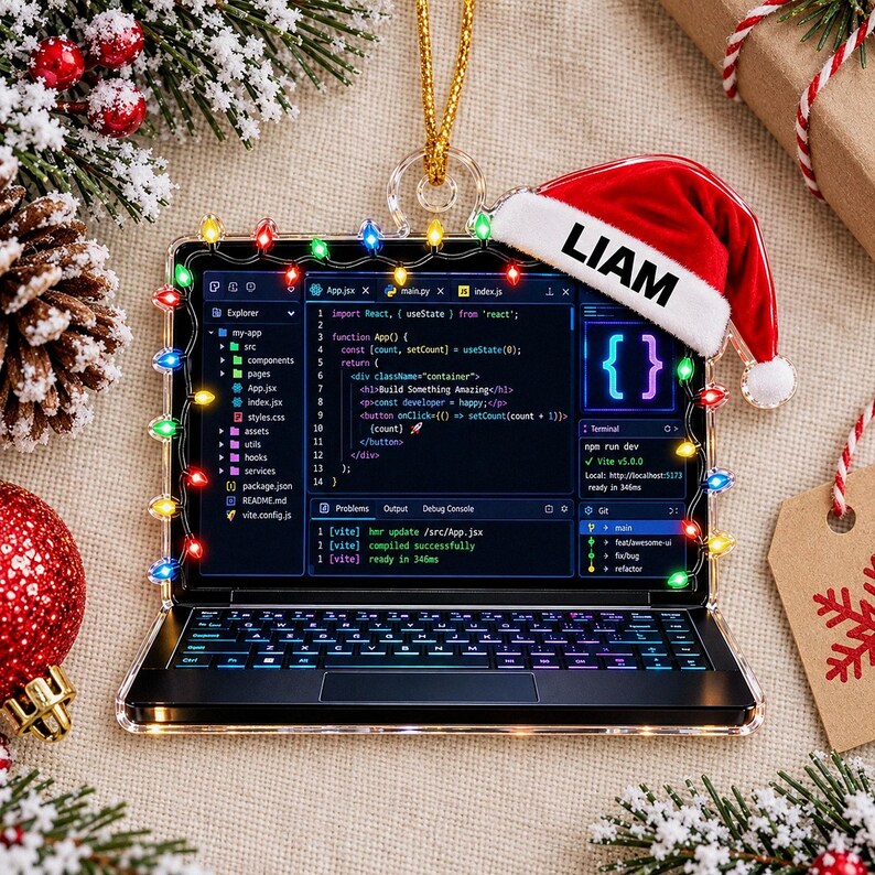May include: A laptop-shaped Christmas ornament featuring a screen displaying code, decorated with colorful string lights around the frame. A red and white Santa hat with the name LIAM printed in black sits on the top right corner of the laptop.