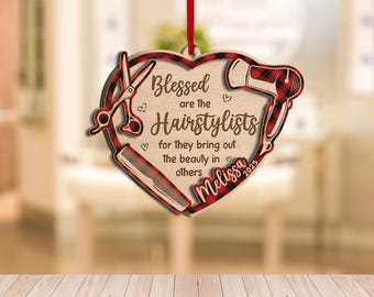 May include: Wooden heart-shaped ornament with a red ribbon for hanging. The ornament features a plaid design and includes a comb, scissors, and a hairdryer. Text reads "Blessed are the Hairstylists for they bring out the beauty in others".
