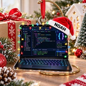 May include: A laptop-shaped Christmas ornament featuring a screen displaying code, a keyboard, and a string of colorful LED lights around the frame. A miniature red and white Santa hat with the name NOAH printed on the brim rests on the top corner.