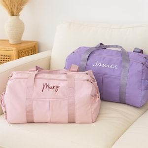 May include: Two personalized duffel bags in soft pastel colors. One is light pink with "Mary" embroidered, the other is lavender with "James" embroidered. Both bags have matching handles and zippers, perfect for travel or gym.