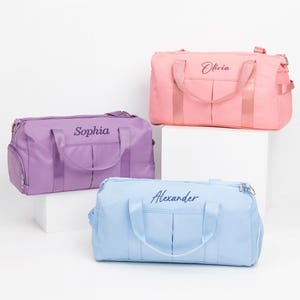 May include: Three personalized duffel bags in pastel colors: lavender, pink, and light blue. Each bag has a name embroidered on the front: "Sophia", "Olivia", and "Alexander". The bags have handles, a shoulder strap, and a front pocket.