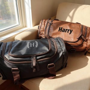 May include: Two personalized duffel bags. One is dark gray with the letter "H" and a laurel wreath, the other is brown with the name "Harry". Both bags have brown accents, zippers, and straps. They are sitting on a beige couch.