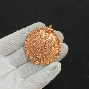 May include: A detailed, rose-gold pendant with an intricate circular design. The pendant features a central floral pattern surrounded by concentric circles and spiral details. The outer edge has a beaded border and a small loop for a chain.