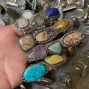 Può includere: Una collezione di anelli con pietre preziose di varie forme e colori, tra cui turchese, ametista e occhio di tigre. Gli anelli hanno montature in metallo color argento con bordi decorativi. Alcuni anelli sono a fuoco, altri sono sullo sfondo.