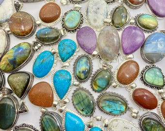 Op de afbeelding: Een verzameling edelsteenhangers met zilverkleurige metalen zettingen. De hangers hebben verschillende vormen en kleuren, waaronder blauwe, groene, bruine en paarse stenen. Sommige hebben ingewikkelde bezel-zettingen, andere eenvoudige lussen.